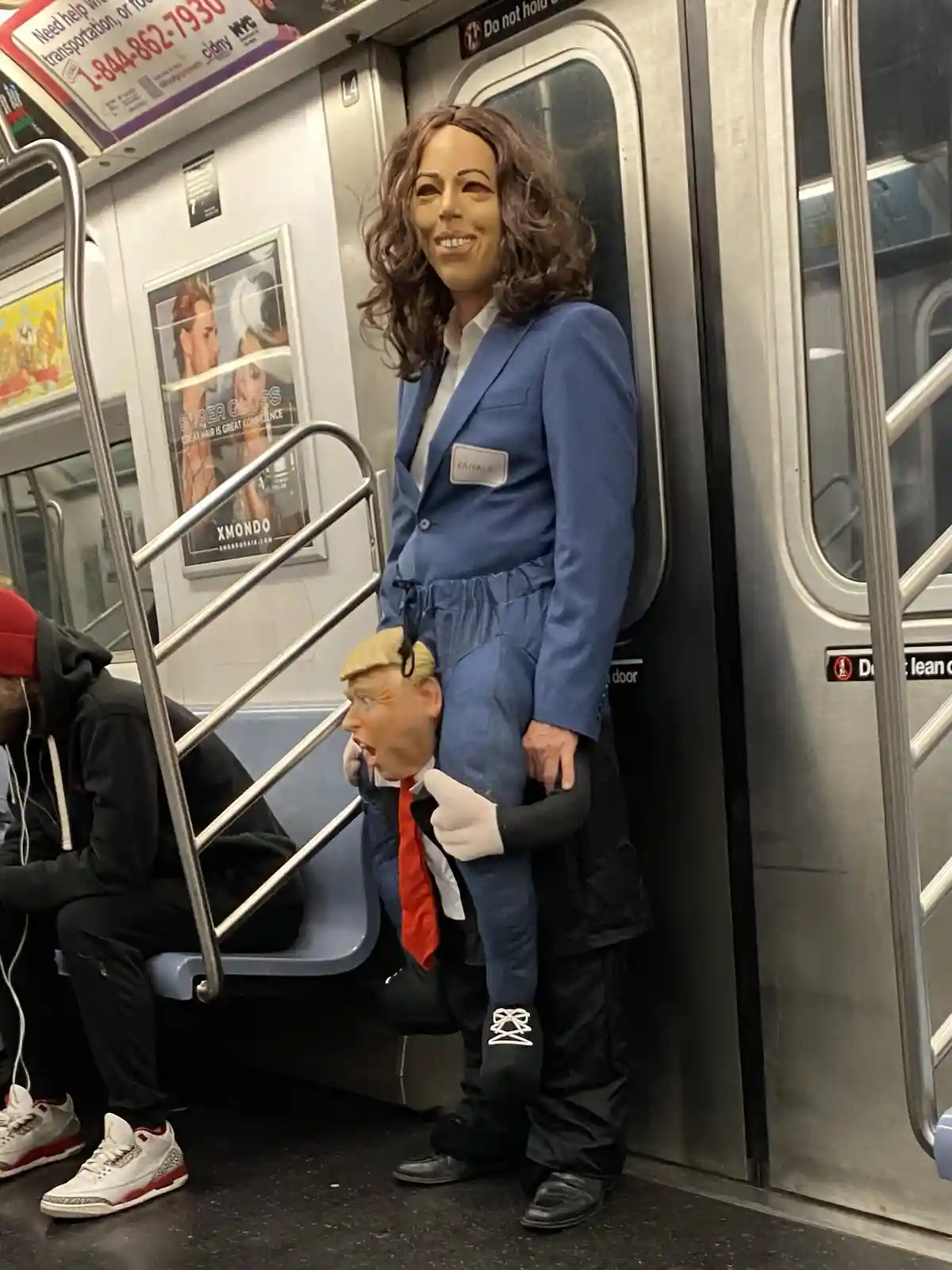 A person in a Kamala Harris costume stands inside a subway car, holding a Donald Trump puppet on their back. The Kamala…
