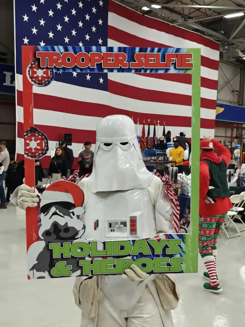 A person in a full white stormtrooper costume stands inside a festive photo frame labeled 'TROOPER SELFIE' and 'HOLIDAYS &…