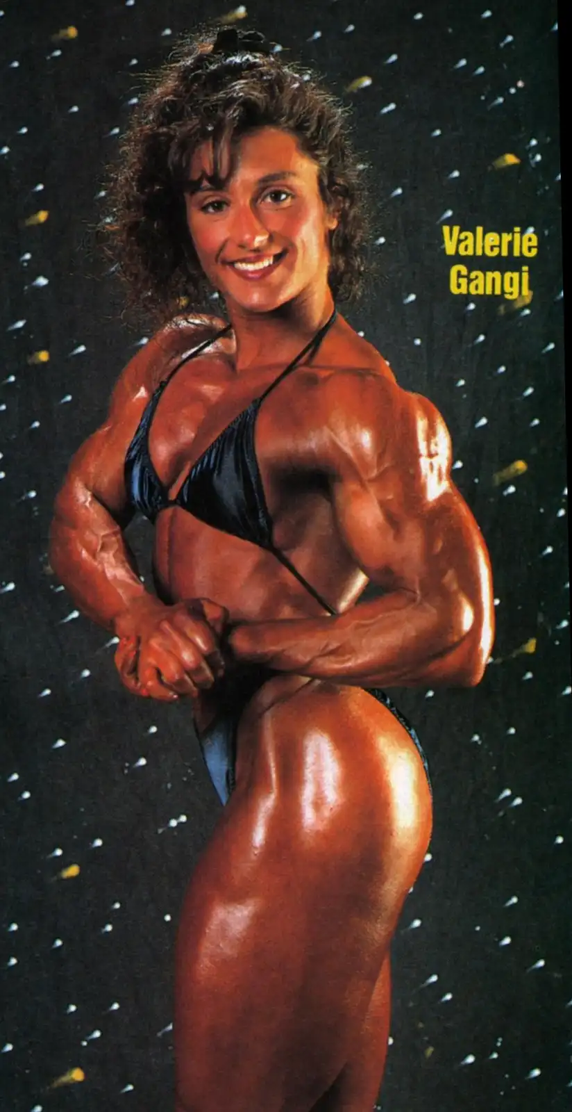 A vintage photo of bodybuilder Valerie Gangi posing in a black bikini against a dark, speckled background, flexing her…
