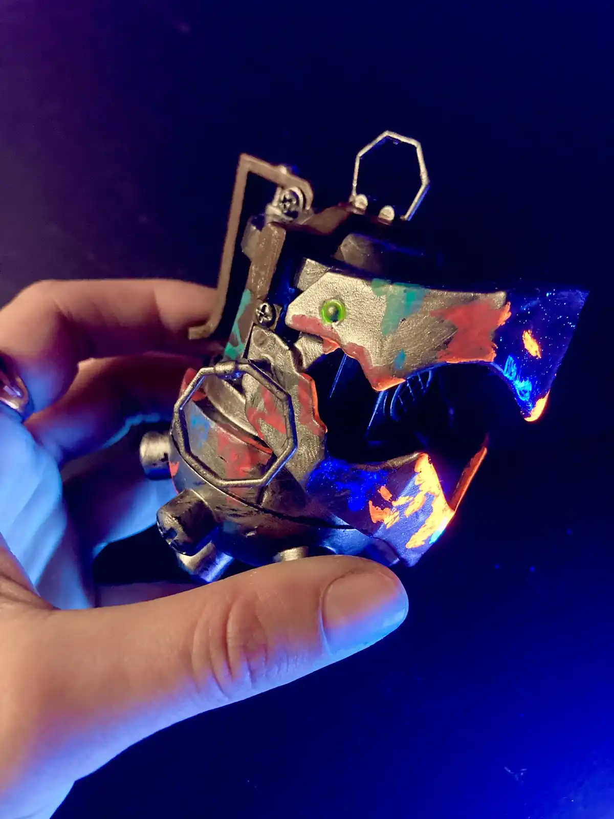 A hand holds a small, custom-painted mechanical-looking object under blacklight, which makes the paint glow with bright…