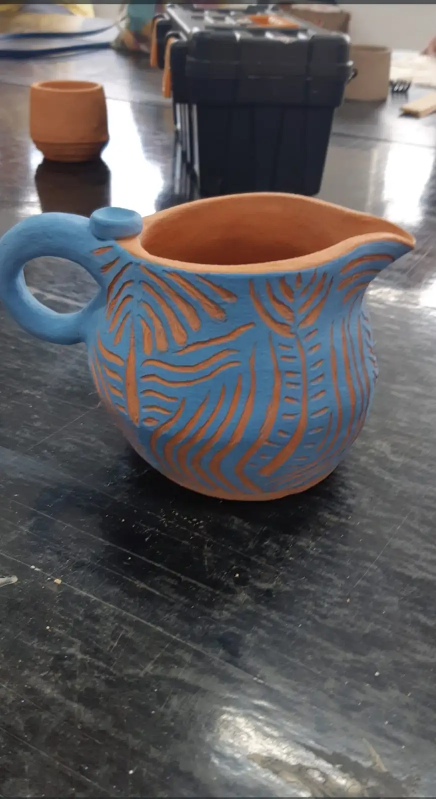 A handmade ceramic pitcher painted blue with orange leaf-like patterns sits on a dark, scratched table. In the background…