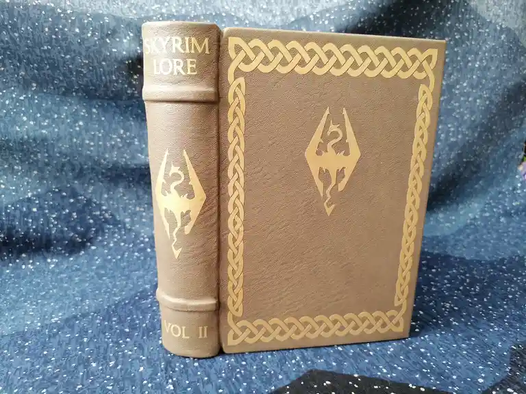 A brown leather-bound book titled 'SKYRIM LORE VOL II' with gold embossed text and a dragon sigil, standing upright on a…