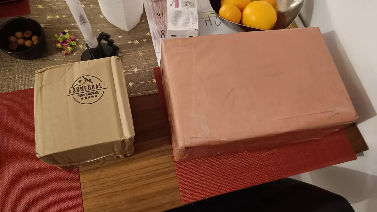 Two wrapped packages sit on a table; the smaller one has a '3DNEURAL 100% HANDMADE' stamp, while the larger one is plain…