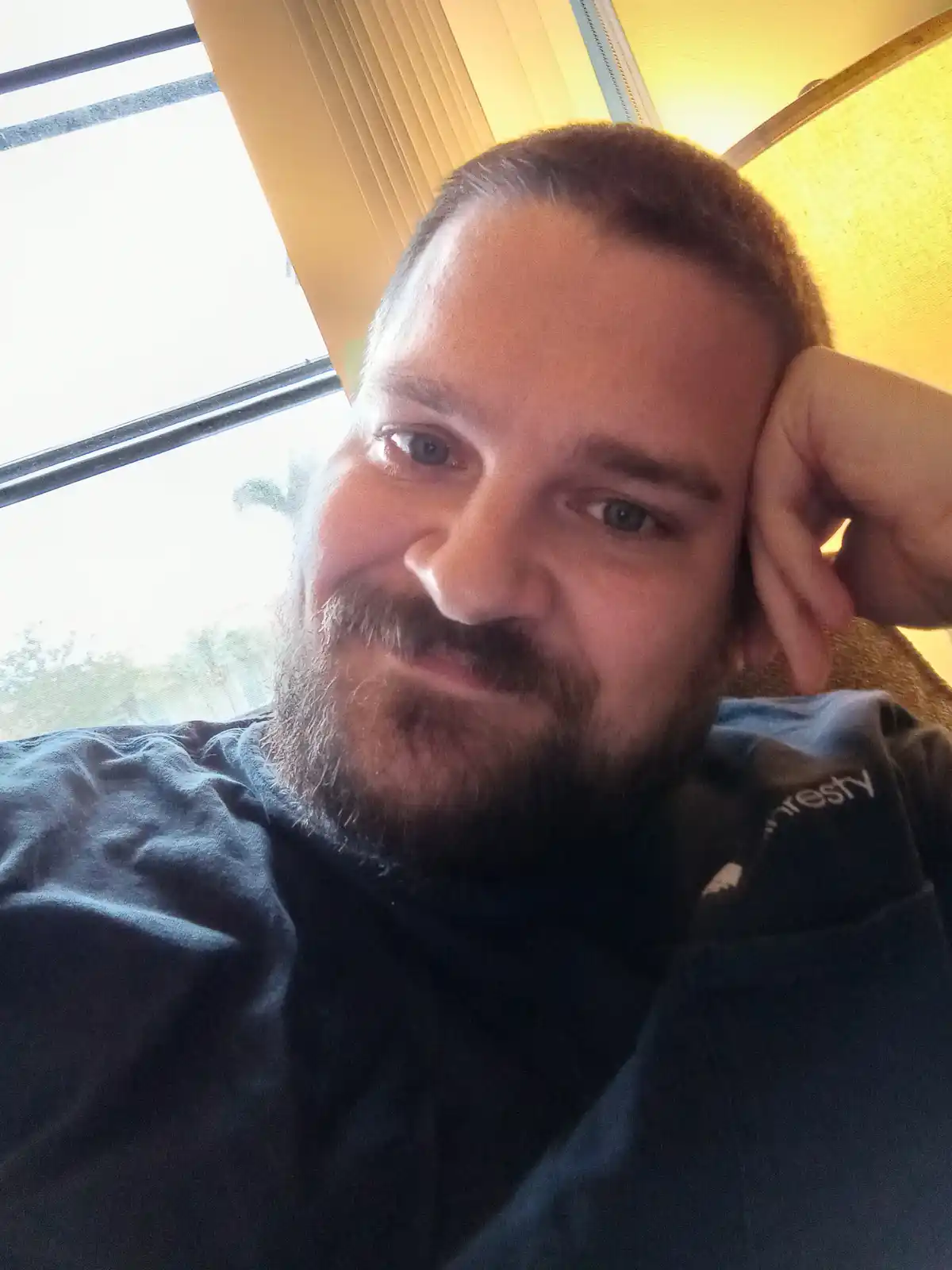 A man with a beard and short hair, wearing a dark blue hoodie, takes a selfie while resting his head on his hand. He is…