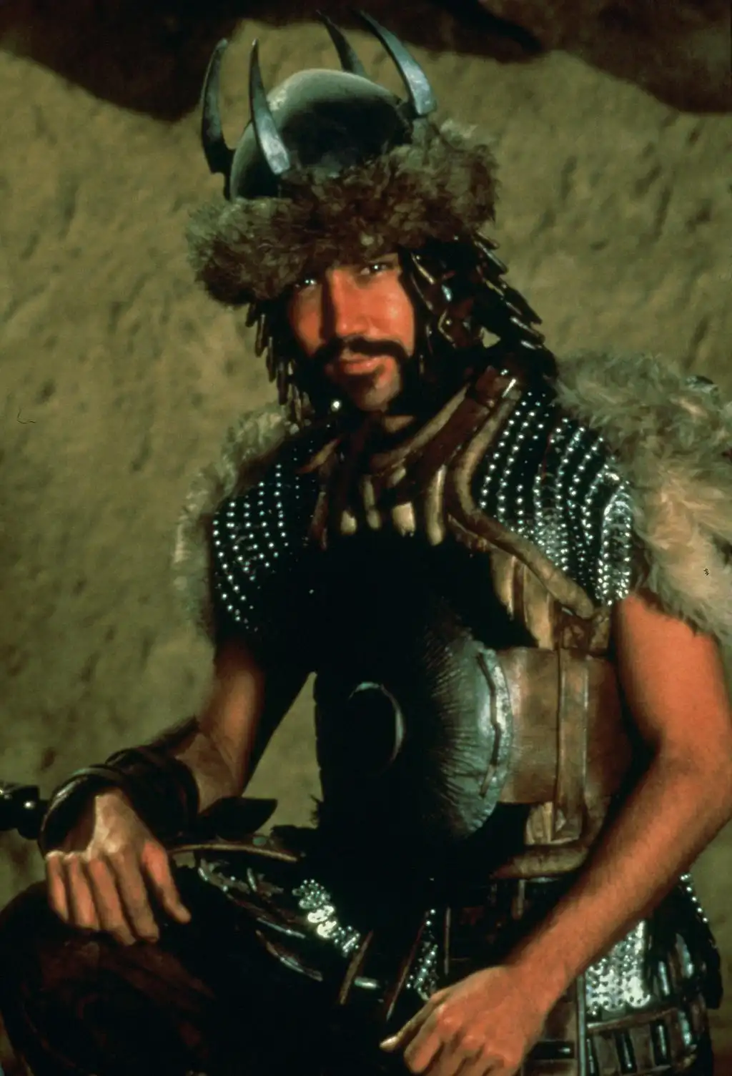 A man in elaborate fantasy armor with a horned helmet and fur trim sits confidently, looking directly at the camera…