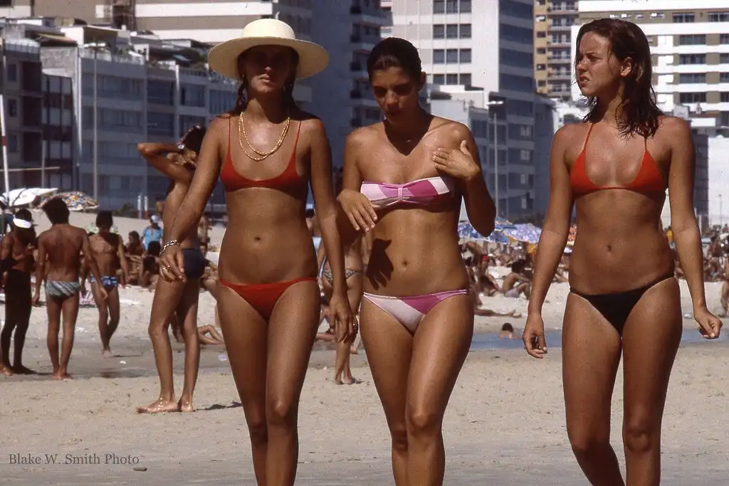 Three women in bikinis walk on a crowded beach with buildings in the background. The woman in the center, wearing a pink and…