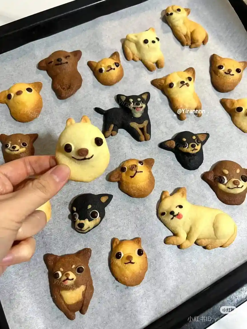 A hand holds up a round, yellow, dog-shaped cookie with a smiling face, while a baking tray below is filled with various…