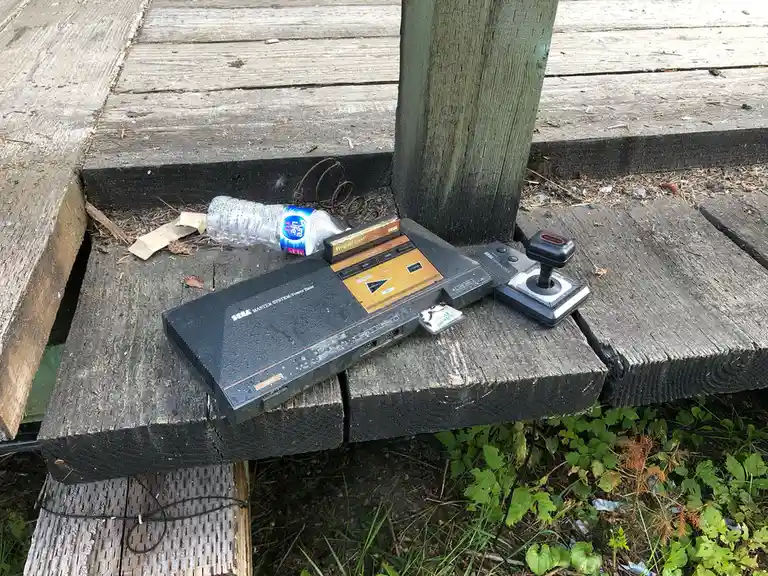 A weathered Sega Master System console and its joystick controller lie abandoned on a dirty wooden deck, next to a discarded…