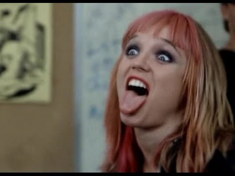 A woman with pinkish-red hair and dramatic makeup stares with wide eyes and an open mouth, her tongue sticking out in a…