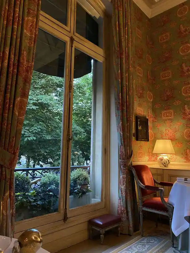 An elegant, ornate room with green and red toile wallpaper, heavy patterned curtains, a red upholstered chair, and a large…