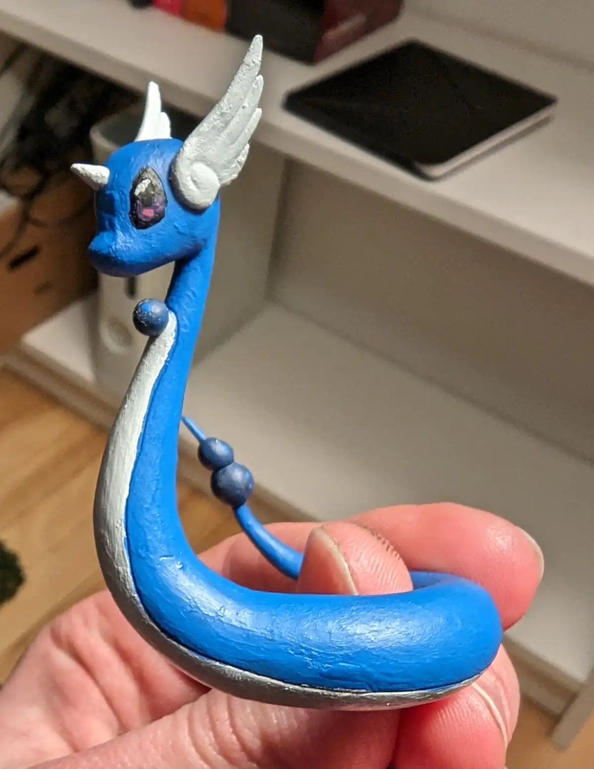 A hand holds a small, blue, hand-painted figurine of a dragon with silver wings and horns, and a purple gem eye. The…