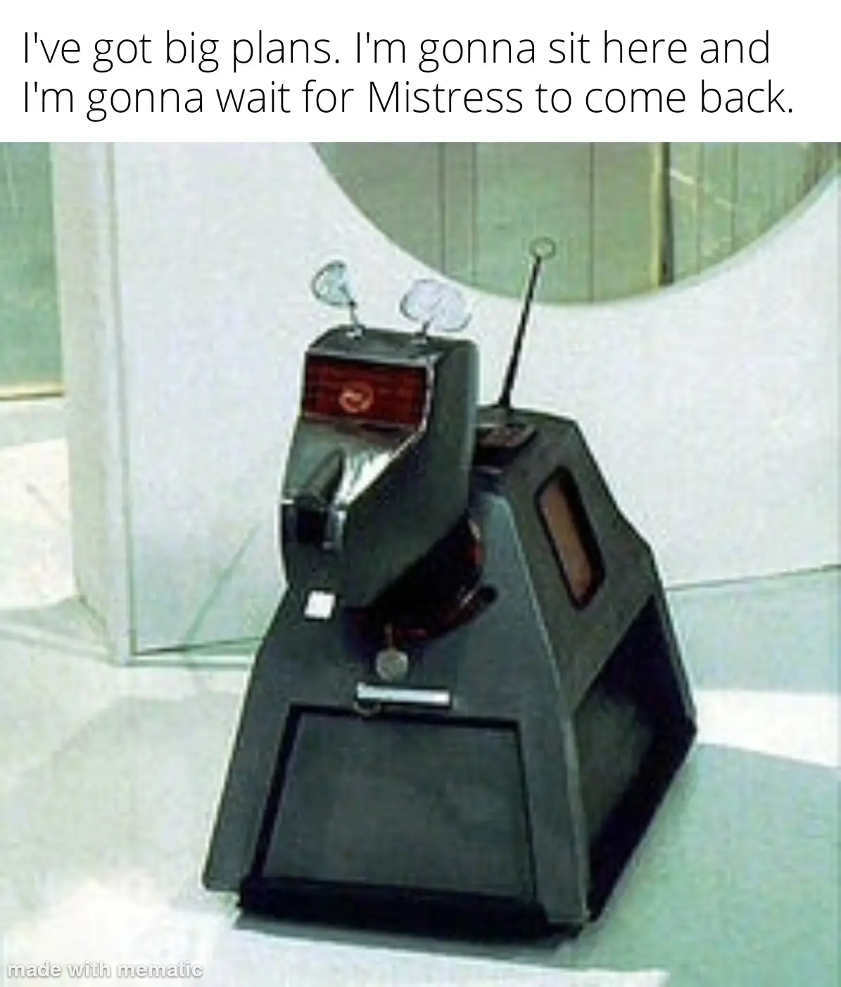 A black, boxy robot with two antennae and a red display panel sits motionless on a white floor in a minimalist room. The…