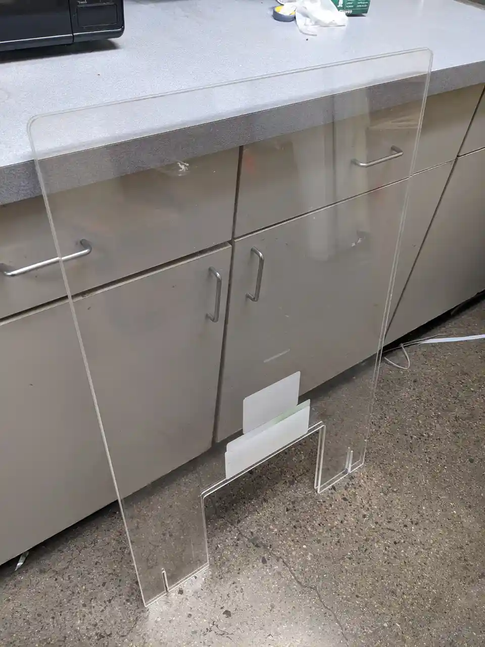 A clear plastic plexiglass barrier stands upright on a speckled concrete floor in front of gray cabinets with silver…