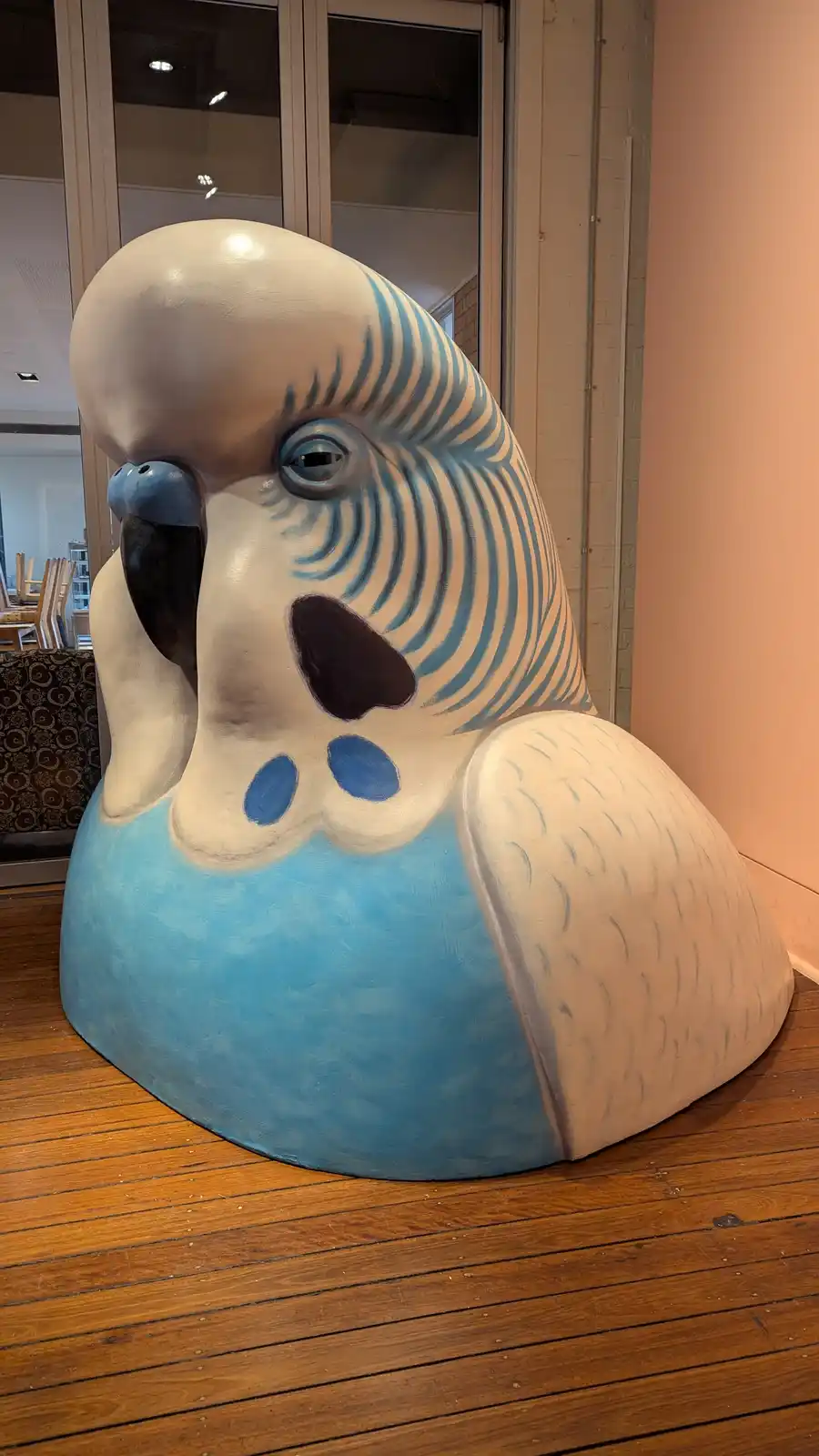 A large, stylized sculpture of a blue and white parrot sits on a wooden floor indoors, its head tilted slightly and its beak…