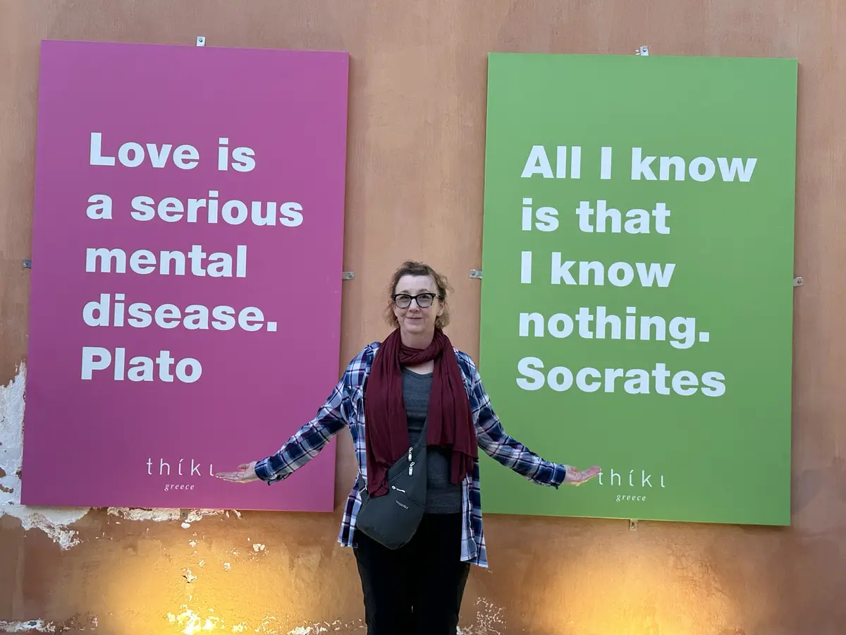 A woman stands between two large signs on a wall. The left pink sign reads 'Love is a serious mental disease. Plato' and the…