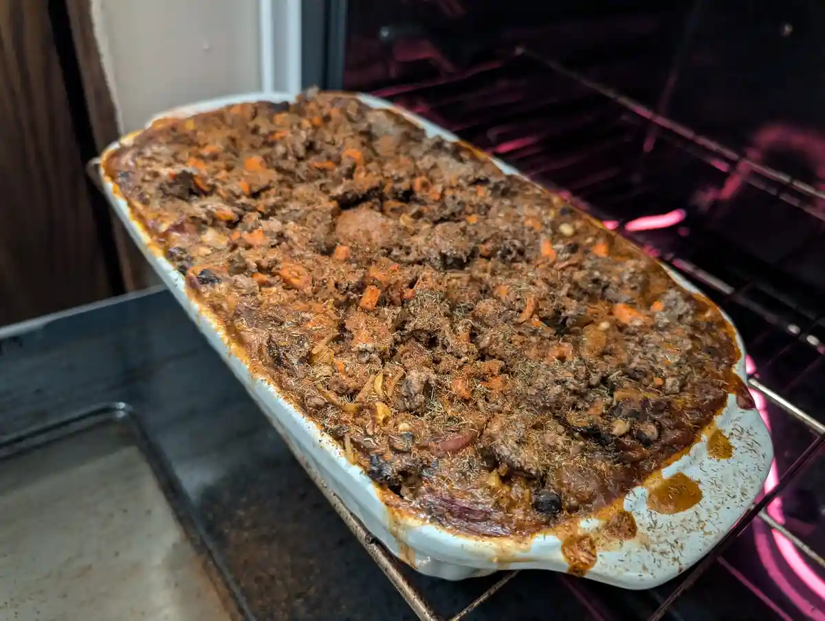 A white ceramic baking dish filled with a brownish casserole containing visible chunks of meat, carrots, and herbs sits…