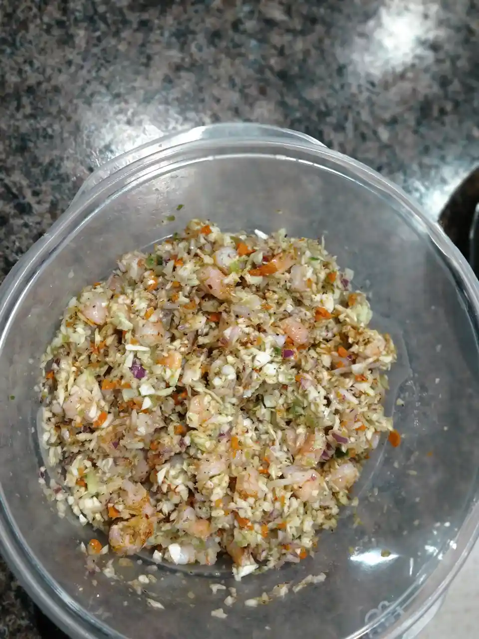 A clear plastic bowl filled with a colorful mixture of finely chopped ingredients including shrimp, cabbage, carrots, and…
