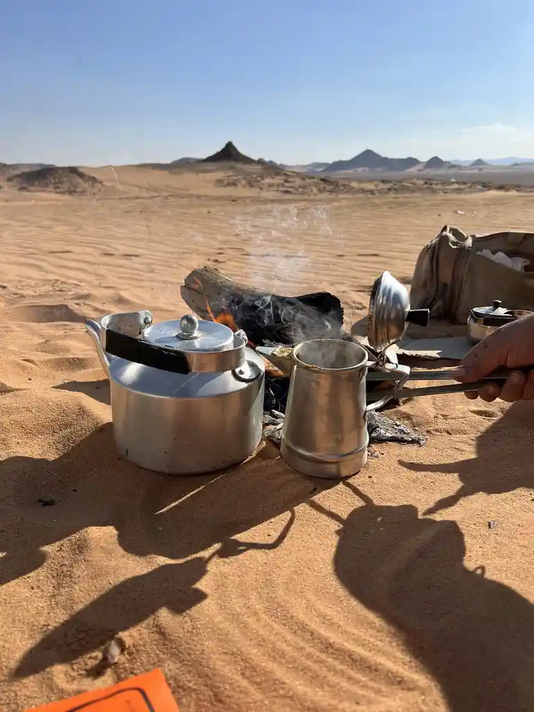 A person's hand holds a metal coffee pot over a small campfire in a vast desert, with a kettle and other camping gear…