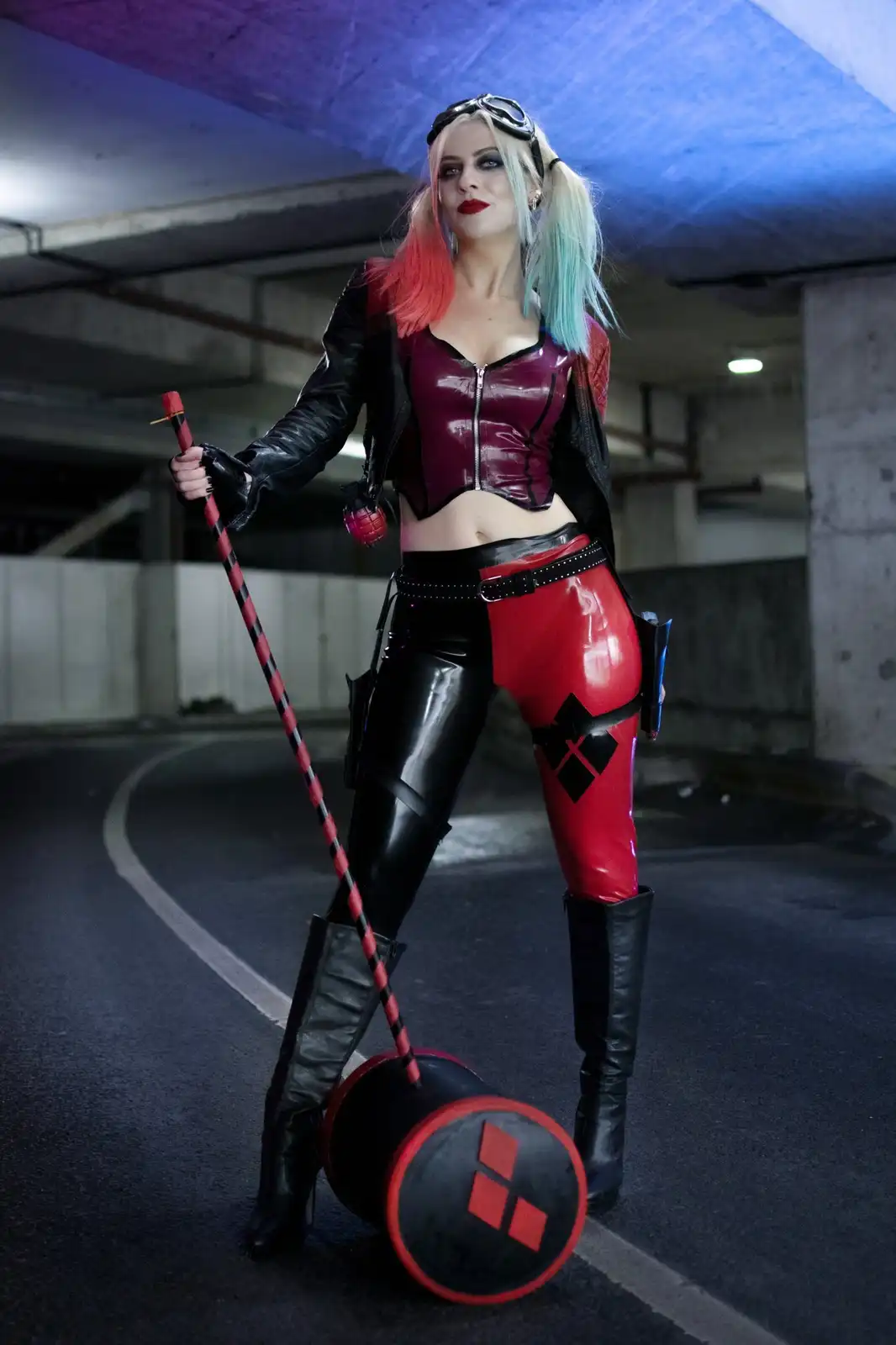 A person in a Harley Quinn cosplay, featuring a red and black latex outfit, pigtails dyed pink and blue, and goggles on…