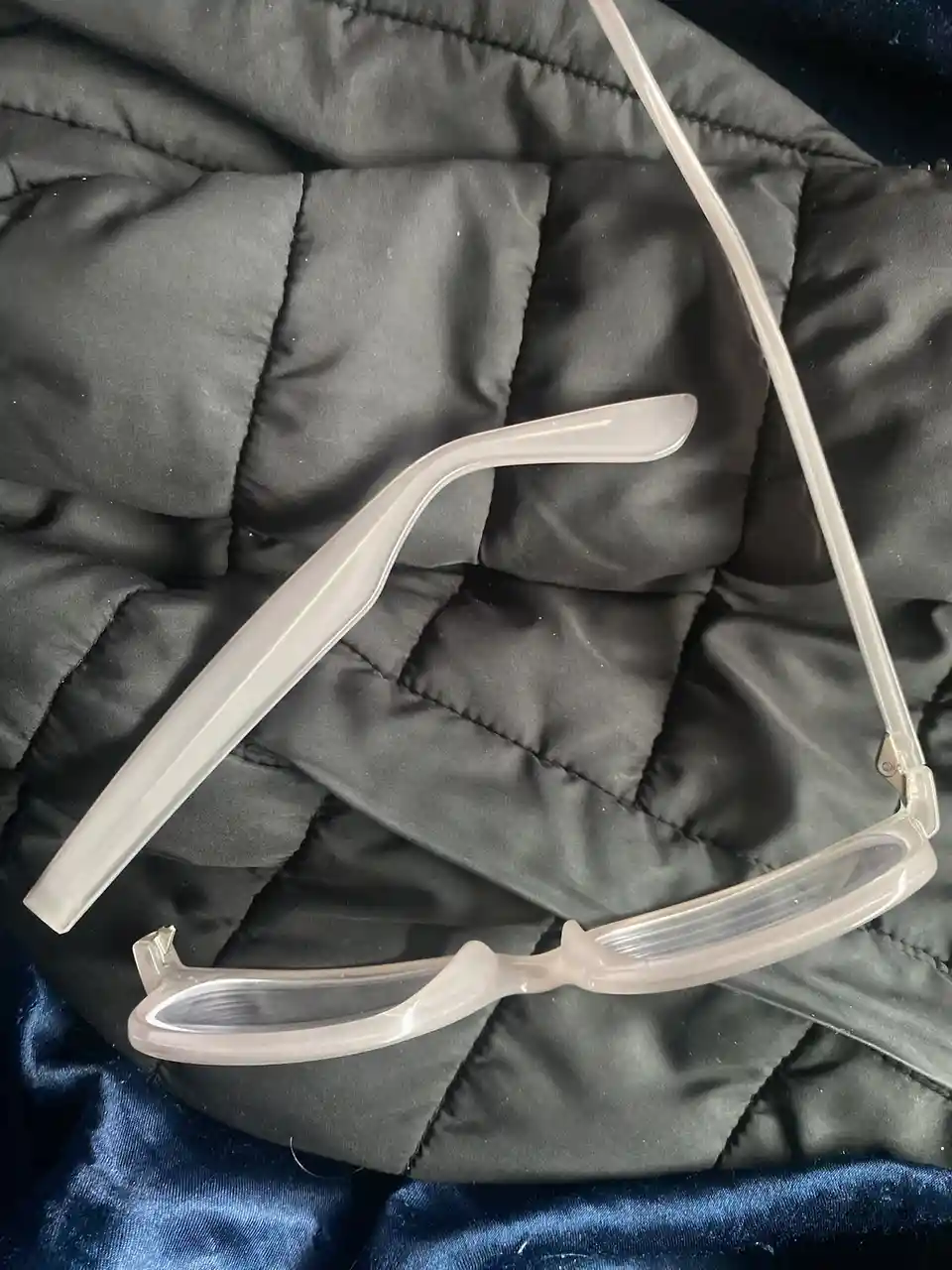 A pair of translucent, light-colored eyeglasses with one broken temple arm lies on a black quilted fabric surface, with a…