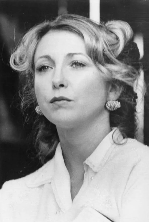 A black-and-white photo of a woman with styled hair and pearl earrings, wearing a light-colored collared shirt, looking off…