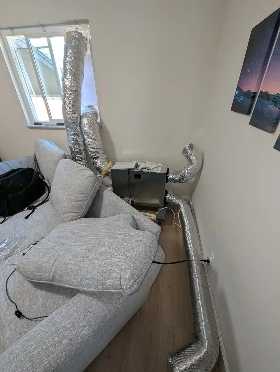 A living room with a gray couch and a window, where a large silver duct is visibly inserted into the window frame, connected…