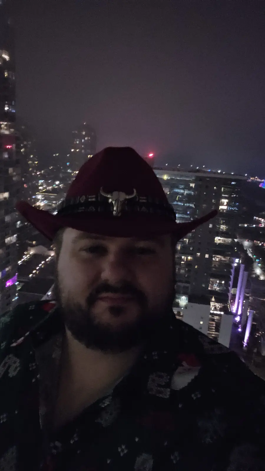 A man with a beard wearing a red cowboy hat with a silver bull skull emblem stands in front of a nighttime cityscape with…