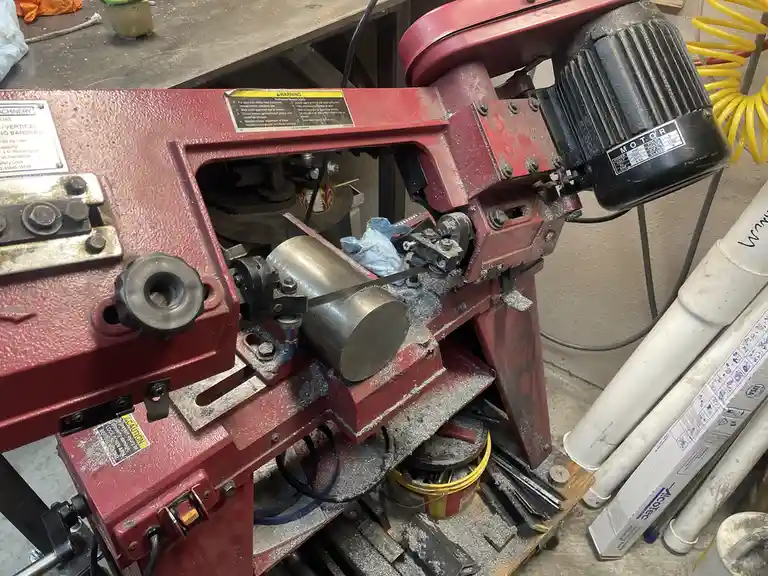 A red vertical bandsaw in a workshop is cutting a metal cylinder, with metal shavings scattered around. The overlaid text…