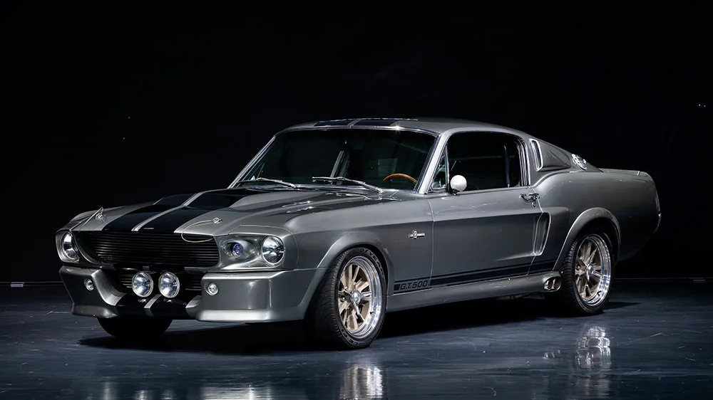 A silver classic American muscle car, resembling a Ford Mustang GT500, is photographed in a dark studio with dramatic…