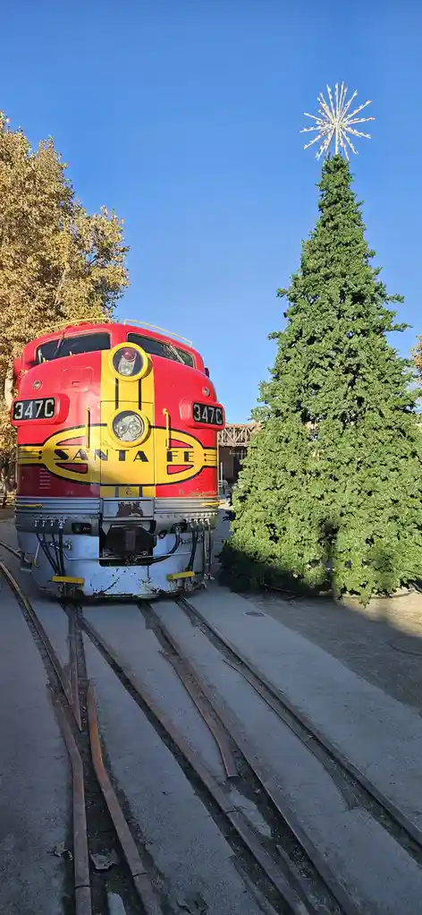 A red and yellow Santa Fe train engine, numbered 347C, is positioned on tracks directly facing a large, decorated Christmas…