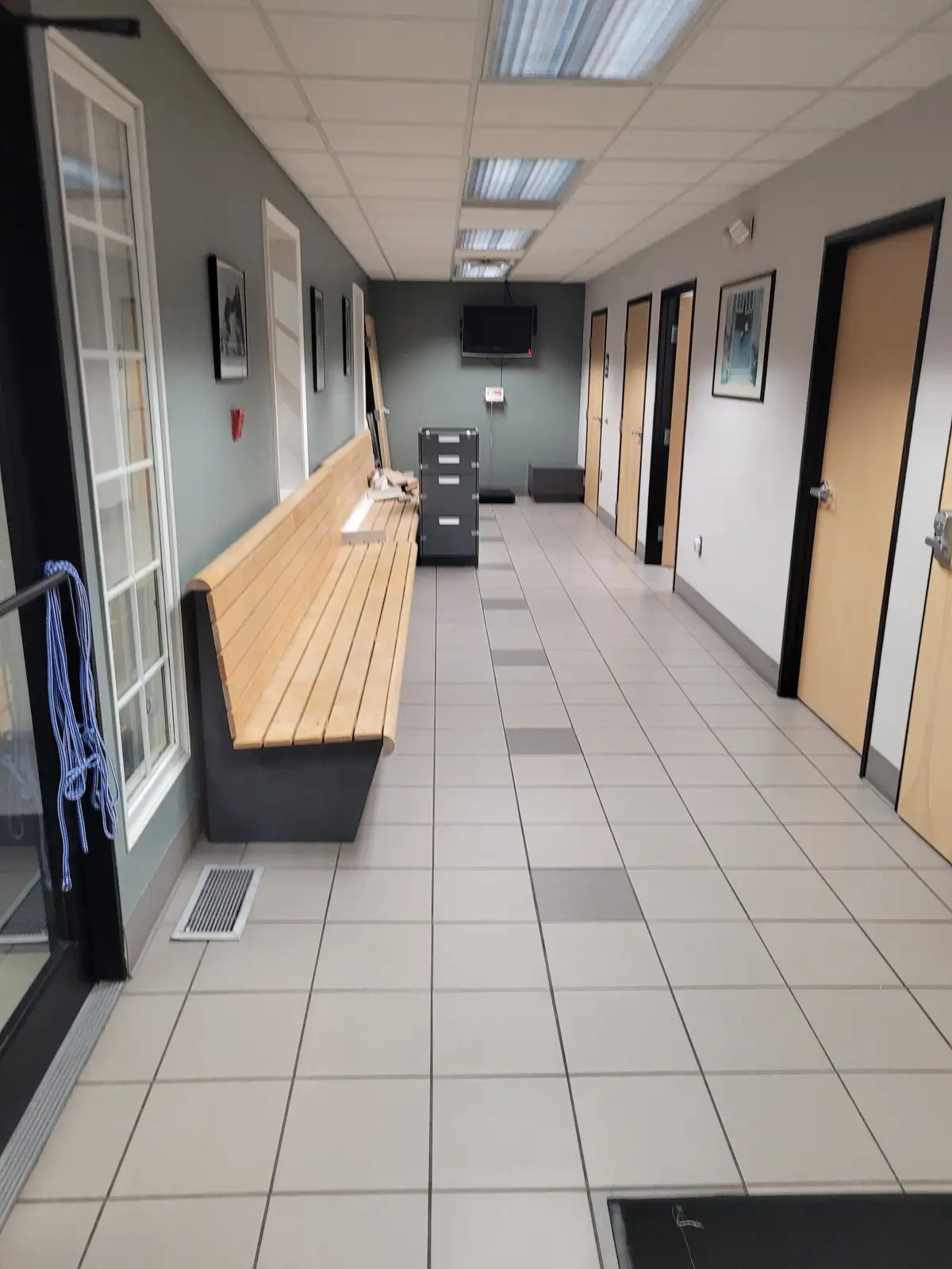 An empty, sterile-looking hallway with tiled floors, wooden benches, and doors on both sides, with a blue rope hanging on a…