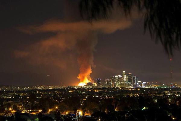 A nighttime cityscape with a large fire and smoke plume rising from the horizon behind illuminated skyscrapers, framed by…
