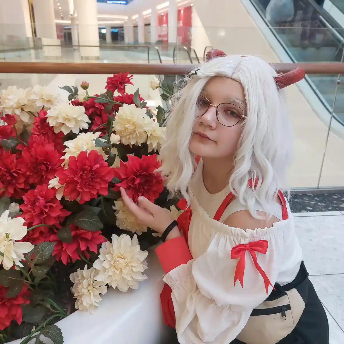 A person with white wavy hair, red devil horns, and glasses is posing beside a large arrangement of red and cream-colored…