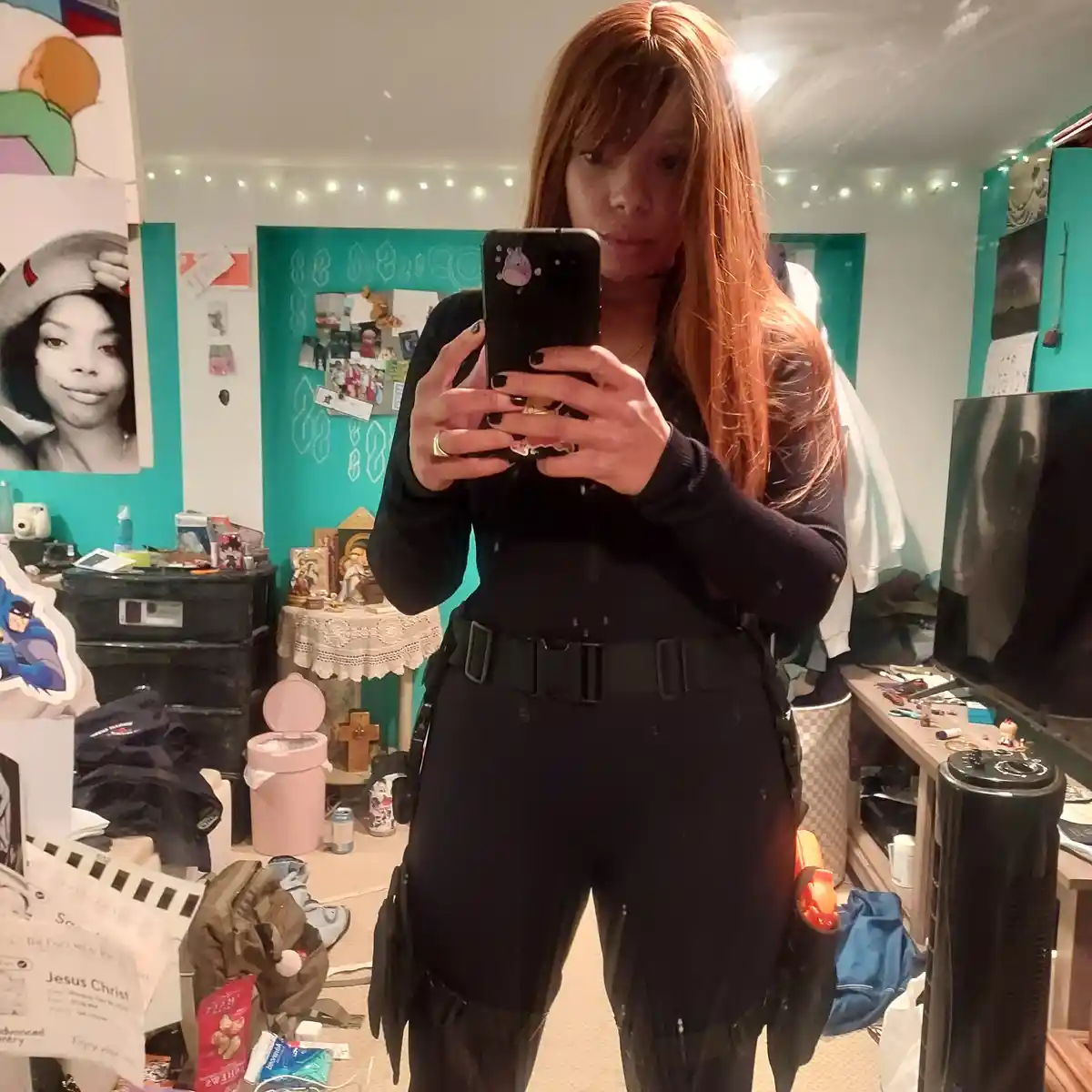 A person with long red hair takes a mirror selfie in a messy bedroom, wearing a black tactical outfit with a belt and…