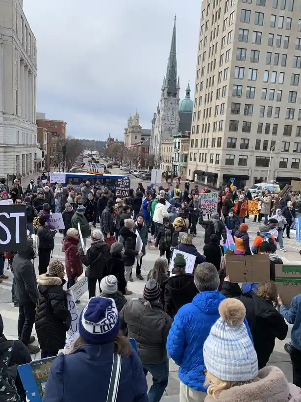 A large crowd of protesters gathers in a city street, holding various signs with political messages. Visible signs include…