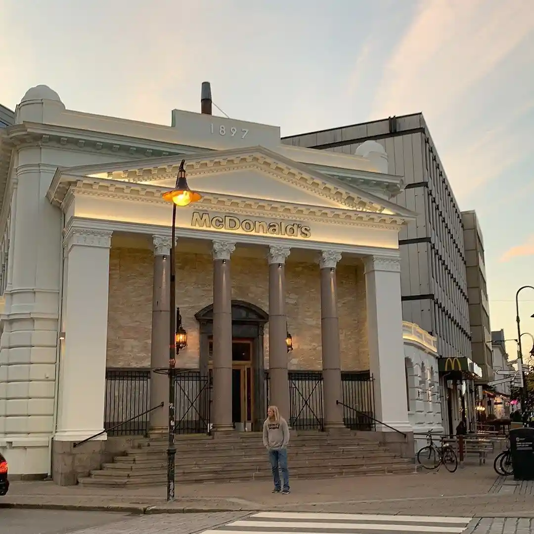 A person stands at the base of grand, classical-style steps leading to a building with columns and a pediment, which has…