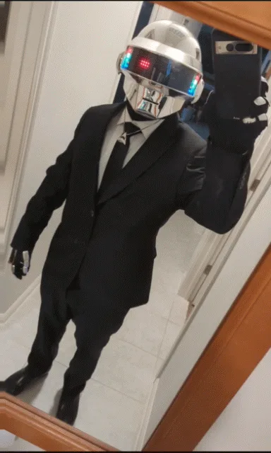 A person in a Daft Punk helmet and black suit takes mirror selfies, holding a phone in one hand and gesturing with the…