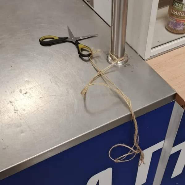 A pair of black and yellow-handled scissors is tied with a frayed string to a metal pole on a stainless steel counter. The…