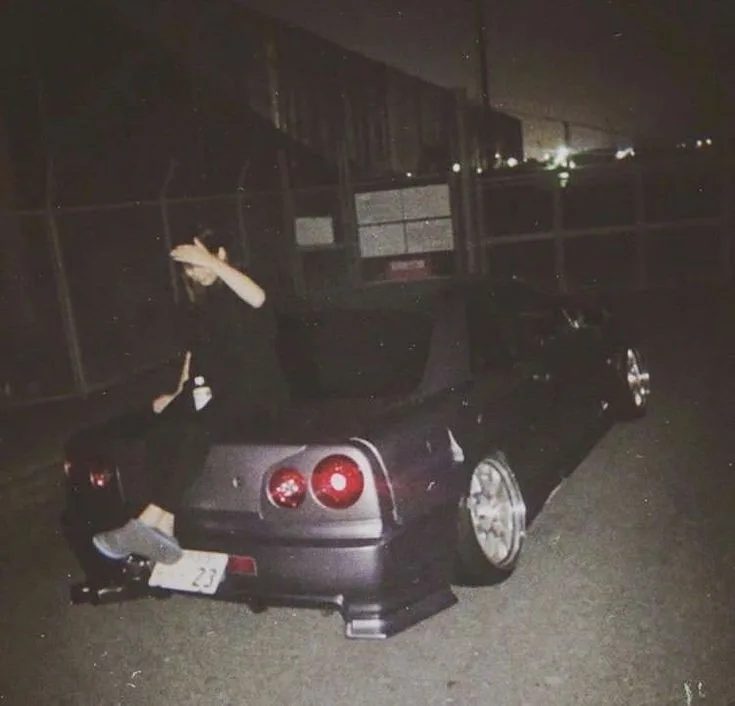 A person sits on the trunk of a dark-colored, modified sports car at night, wearing dark clothing and a cap, with one hand…