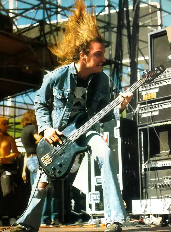 A man with long, wild, spiky blonde hair is energetically playing a bass guitar on stage, wearing a denim jacket and ripped…