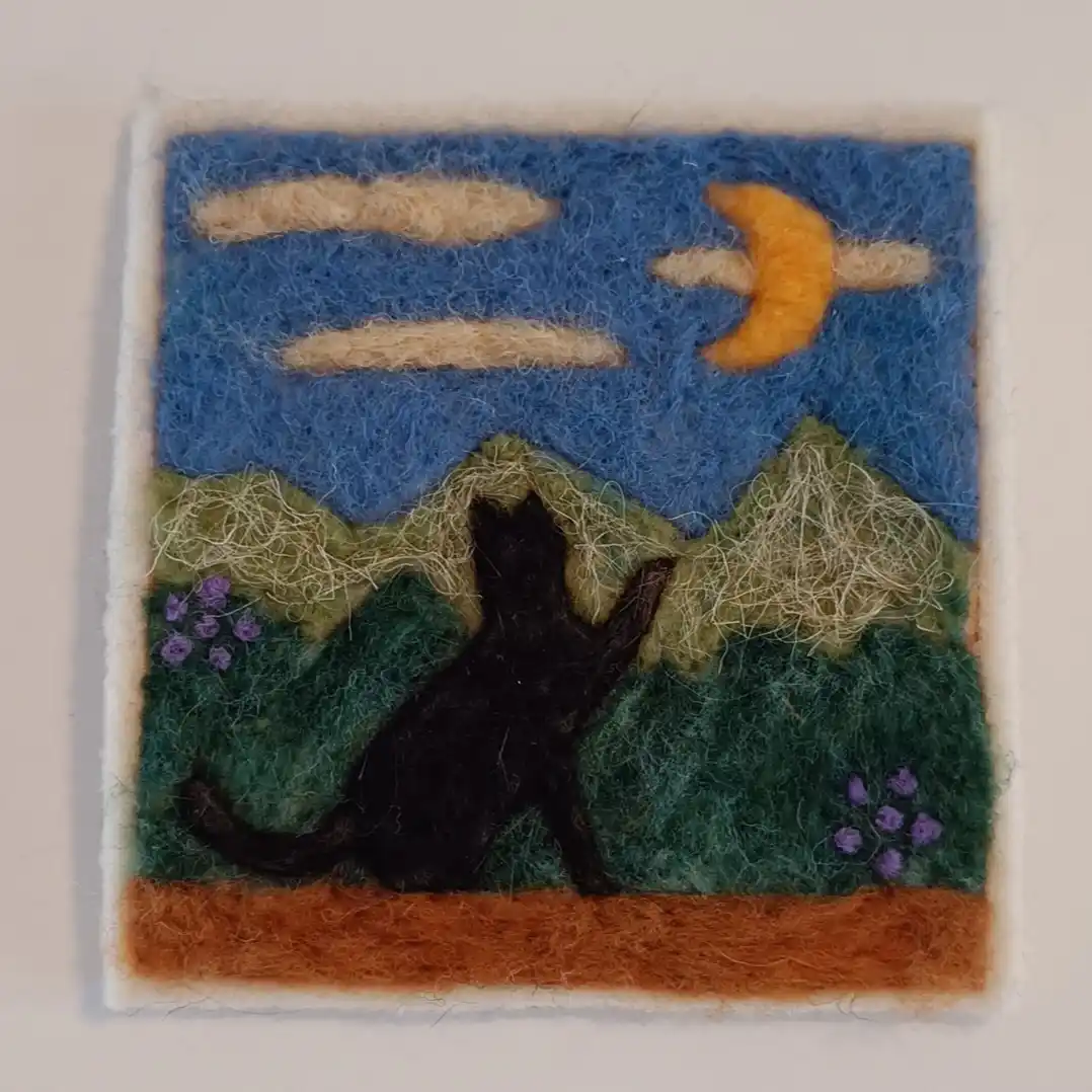 A felt art piece depicts a black cat sitting on a brown ground, looking up at a blue sky with a crescent moon and clouds…