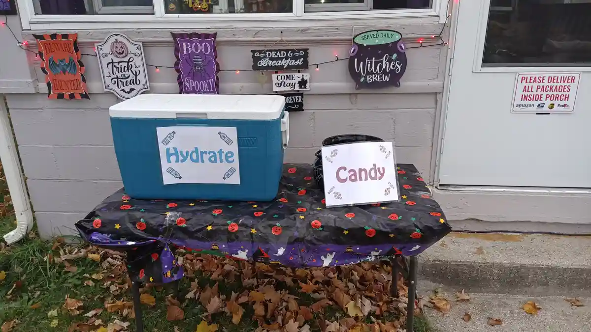 A Halloween-themed outdoor table with a blue cooler labeled 'Hydrate' and a candy bowl labeled 'Candy', set against a house…