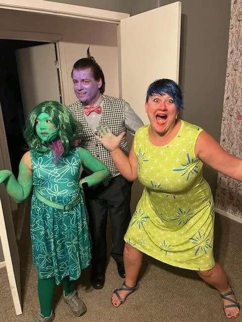 Three people in costumes stand in a doorway; the person on the left has green face paint and wig, the center person has…