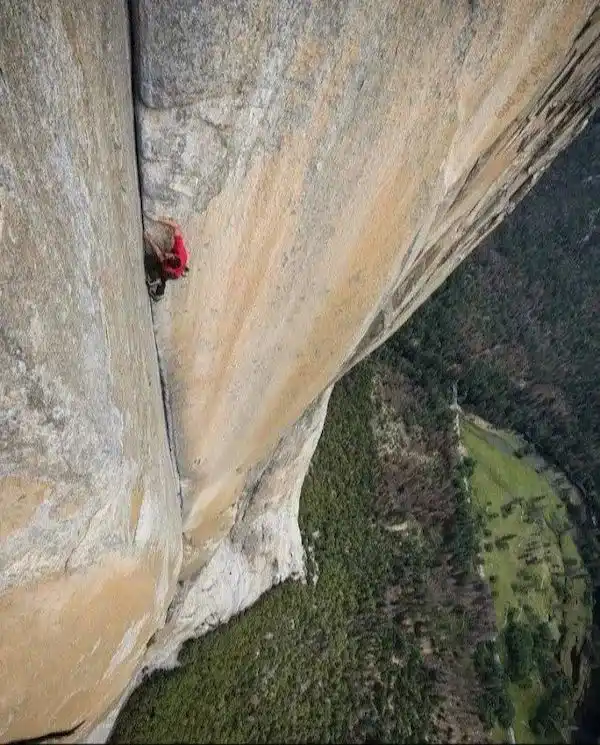 A climber in a red shirt is clinging to a narrow vertical crack in a massive, sheer rock face, with a vast green valley far…