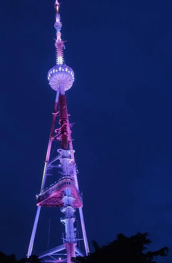 A tall, illuminated broadcast tower stands against a dark blue night sky, its structure lit with purple and white lights…