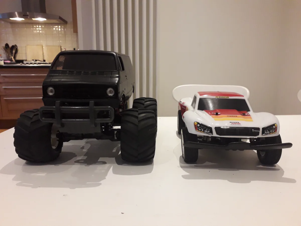 Two remote-controlled monster trucks sit side-by-side on a white surface in a kitchen. The truck on the left is black with…